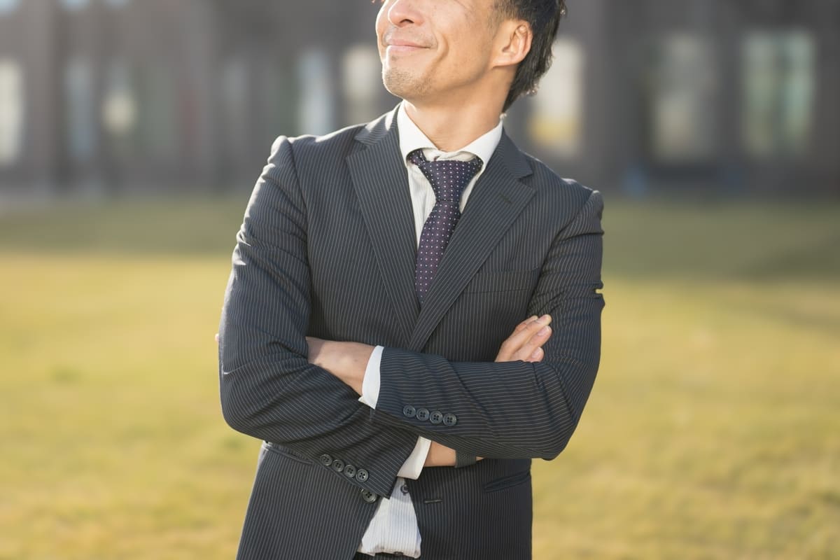 スーツ姿で腕を組み、満足そうに笑みを浮かべる男性会社員。背景は都市の建物で、不動産投資への第一歩を踏み出した自信と決意を象徴する構図。